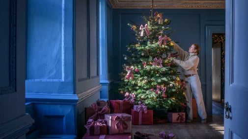 A large decorated Christmas tree visible through the doorway of the Great Chamber in Avebury Manor, with a member of staff applying finishing touches.